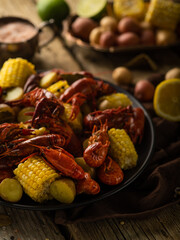 The photo shows a close-up of a bright colorful dish - boiled crayfish with pieces of boiled corn. Also in the photo there is a lemon and many different ingredients in the background. Rustic style.