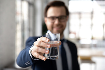Happy businessman holding glass of fresh filtered cold water, toasting at camera. Male employee drinking still mineral water during break, keeping hydration aqua balance. Close up of hand