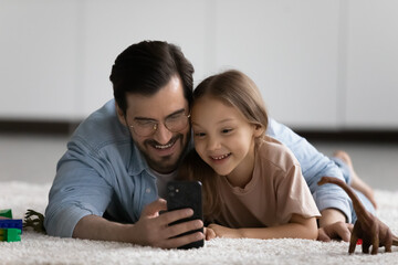 Loving happy young Caucasian father and little daughter lie on floor at home laugh using smartphone together. Caring smiling dad and small girl child talk on webcam video call on cellphone.