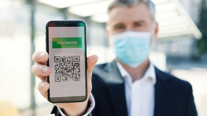 Close up of middle-aged Caucasian man traveller standing outdoor on street in medical mask holding smartphone showing QR code with covid-19 vaccination digital passport, vaccination certificate
