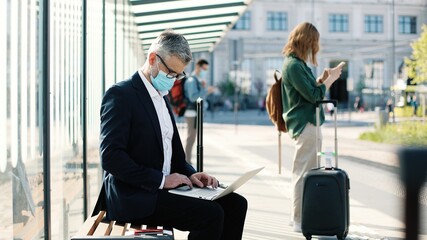 Side view of Caucasian middle-aged businessman in suit and medical mask sitting on bus stop and typing on laptop outdoors. Business trip during covid quarantine. People waiting on bus on street