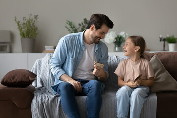 Smiling young Caucasian father and small daughter relax at home play with wooden toy guns in war. Happy dad and little girl child feel playful have fun engaged in funny game activity together.