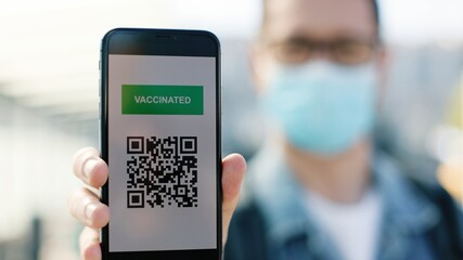 Close up of Caucasian handsome man tourist in medical mask holding smartphone with health vaccination passport for Covid-19. COVID vaccine certificate. Safe travel measures QR code on mobile phone app