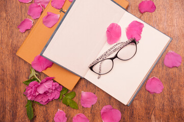 Open book, glasses for reading and rosehip flower petals on a wooden table
