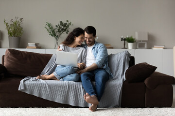 Fototapeta na wymiar Happy millennial multiethnic man and woman relax on couch at home using laptop together. Smiling young multiracial couple rest on sofa in living room have fun browsing internet on computer.