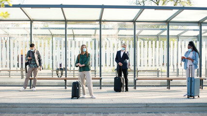 Obraz premium Diverse mixed-race people in masks standing at bus stop or train station with suitcases keeping social distance during coronavirus pandemic. Man speaking on smartphone, young guy texting on cellphone