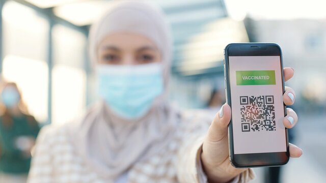 Muslim women in medical mask showing an International Vaccination Certificate COVID-19 QR code on smartphone. Close up concept. Vaccinated person using digital health passport app in mobile phone