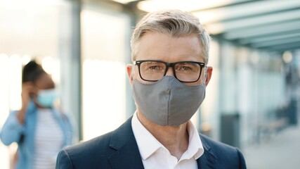 Close up of joyful adult Caucasian man in glasses wearing mask on face standing in town on street and looking at camera. Happy handsome male in good mood outdoors at bus stop