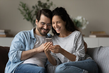 Obraz premium Smiling young multiethnic couple relax at home look at cellphone screen browse wireless interne together. Happy millennial multiracial man and woman use smartphone talk on video webcam call.