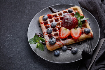 Delicious waffles with berries, mint and sweet sauce on black plate on grey background. Top view....