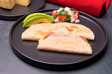 ham and cheese quesadillas served with avocado slices and pico de gallo sauce on a black plate. in horizontal view