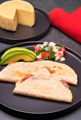 ham and cheese quesadillas served with avocado slices and pico de gallo sauce on a black plate. in portrait view