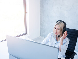 A female office worker sits at a desk and uses a computer and headphones.