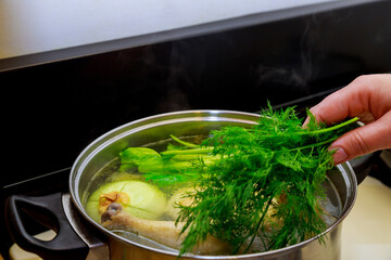 Woman put dill in the chicken broth with whole chicken and onion in pot.