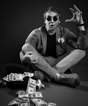 Blind beggar, pauper man in jacket, jeans and shoes sits cross legged on floor by upside down hat full of dollars cash and tells thrilling stories over dark background. Black and white