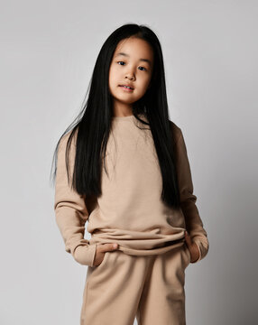 Portrait of smiling asian kid girl in green casual sportswear pants, hoodie, jersey and sneakers standing with hands in pockets ogf her pants over gray background