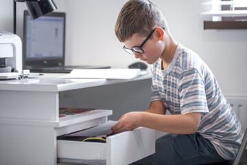Obraz premium An elementary school student sits at a table, takes something out of a drawer.