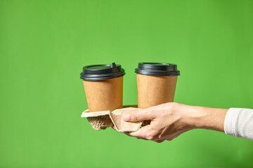 Hands holding two cups brown paper with black lid. Two coffee special offer or promo. Hands holding two cups on green background. Tea or Coffee to go. Brown paper cup with black lid.