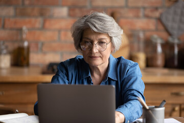 Serious focused middle-aged woman in glasses working on laptop seated at table in kitchen, lead...