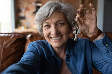 Happy mature smiling woman holding camera showing bunch of keys from new own house, close up. Bank...