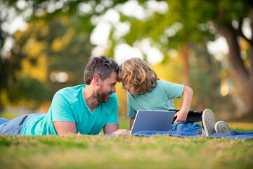 Happy father with son use laptop. Dad and school boy child looking screen and tablet, watching video lesson, sitting on grass. Family weekend. Video call of a grandson to grandparents.