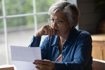 Older female sit in kitchen wear glasses reading letter received information feels upset looking...