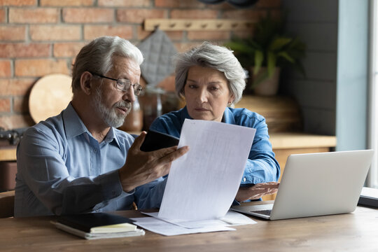 Focused worried grey haired elderly spouses sit in kitchen with laptop on table reading financial papers documents, checking bills looking concerned, planning budget expenses, discussing debt concept