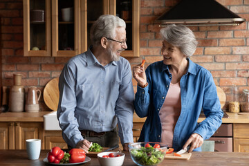 Older couple cooking together vegetarian food in kitchen. Mature spouses look at each other with...