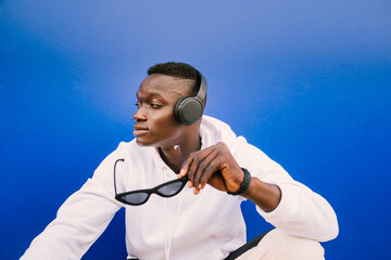 Young afro American black man wearing a white sweatshirt, sunglasses and headphones on a blue wall looking to the side. Modern look