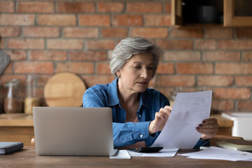 Middle-aged woman sit in kitchen hold sheet document paper read letter check notice rates, pay bills use online e-bank app. Finance management, examining bank statement, family budget planning concept