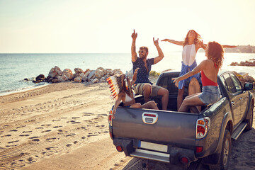 A group of handsome models is sitting in the back of a car by the sea and having fun while listening to the music. Summer, sea, vacation, friendship