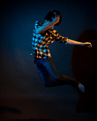 Fototapeta na wymiar A young man in a plaid shirt jumps on a dark background illuminated by blue and yellow light.