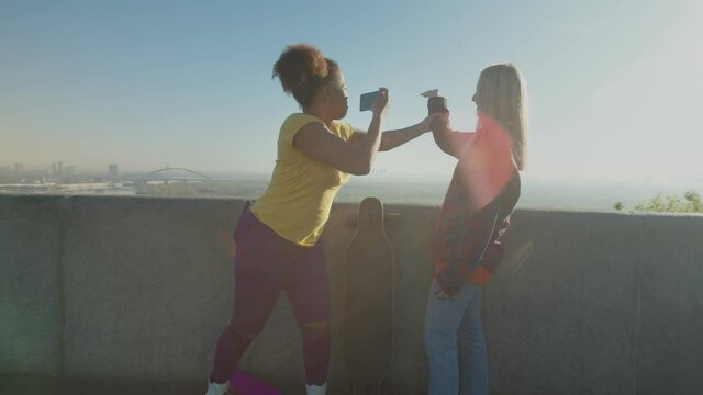 Carefree mischievous diverse multiracial women skateboarders taking a break from skateboarding, having fun, making funny selfie shot on smart phone while relaxing at observation deck at daybreak.