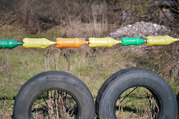Fototapeta premium Plastic colored bottles hang in a row over black car tires against the backdrop of spring nature and piles of rubble in the background.