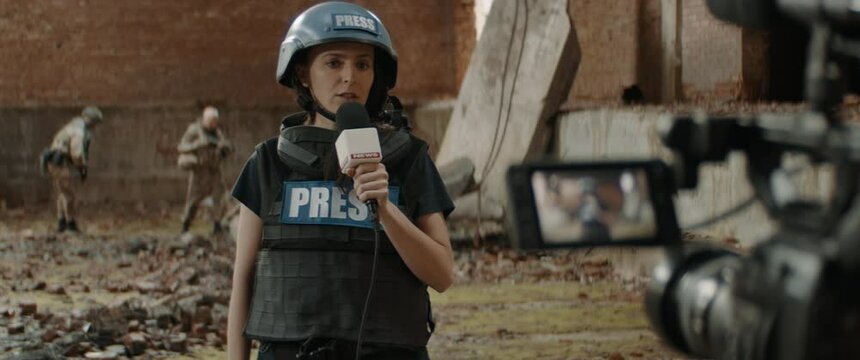 Behind the scenes of female war journalist correspondent wearing bulletproof vest and helmet reporting live near destroyed building, military personnel in the background