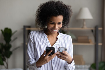 Happy millennial African American woman use two modern smartphones synchronize devices. Smiling...
