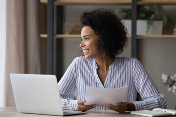 Happy millennial mixed race female employee work on computer at home office look in distance thinking dreaming. Smiling successful young African American woman busy with paperwork using laptop.