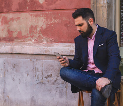 Serious male entrepreneur in elegant suit standing on street and surfing internet via smartphone