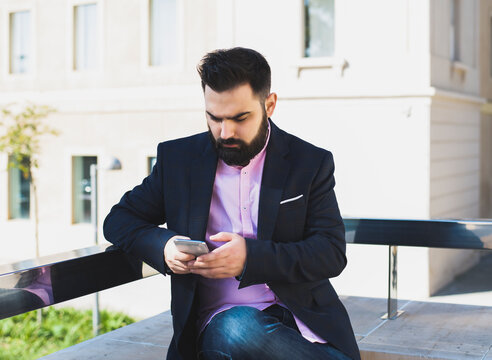 Bearded young man dressed in suit uses his smartphone on the street