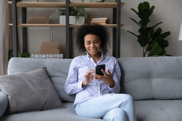 Happy young African American woman relax on couch in living room browse wireless internet on...