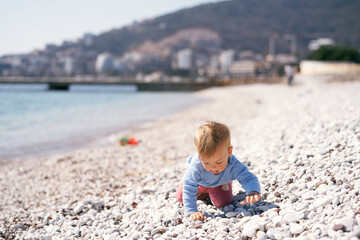 Pensive kid crawls along a pebble beach, sticking out his tongue, by the sea against the background of mountains