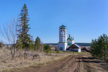 Fototapeta na wymiar Ancient rural church with a bell tower and golden domes in spring. View of the old white church in the village of Klyuchi (Perm Territory, Russia) with a dirt dirt road and trees against the blue sky.