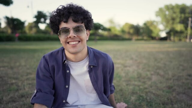 Pov shot of trendy young gen z curly guy with sun glasses talking to his followers in a clip for social network sitting on a city park. Vlogger or internet entrepreneur using video call technology