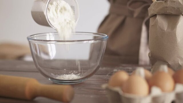 Kneading the dough. The chef pours the flour from a measuring glass into a transparent bowl. The process of preparing bakery products. Products for the dough on the table. Flour paper bag, egg, rollin