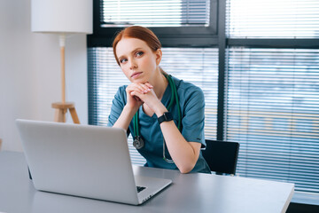 Portrait of thoughtful young female doctor in blue green medical uniform sitting at desk with laptop on background of window in hospital office of medic clinic, looking down. - Powered by Adobe