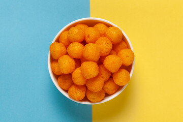 Close up of Cheese Potato Puff Ball Snacks, tangy orange color, Popular Ready to eat crunchy and puffed snacks,  salty in white ceramic bowl over blue yellow background