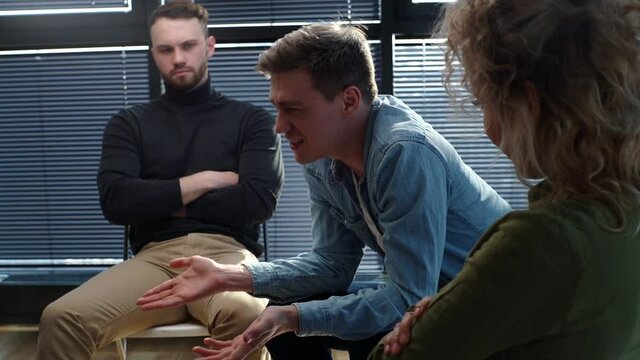 Close-up of sad smiling young man sharing problem sitting in circle during group interpersonal therapy session. Tracking shot of depressed male telling sad story of mental problem to other patients.