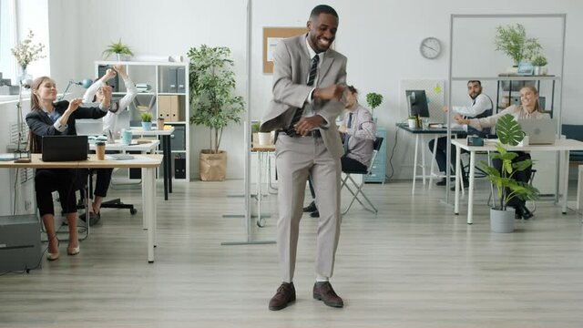 Portrait of enthusiastic African American employee in formalwear dancing in workplace while colleagues men and women are having fun at desks indoors