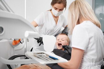 Boy sitting in dental chair while two female dentists checking kid teeth. Woman stomatologist examining little boy teeth with dental instrument while assistant in medical mask pointing at mirror.