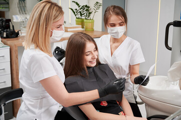 Horizontal snapshot of female dentist and assistant explaining X-ray report to patient in clinic. Young girl in dentist's chair, wearing bib, listening to doctor. Dentistry concept